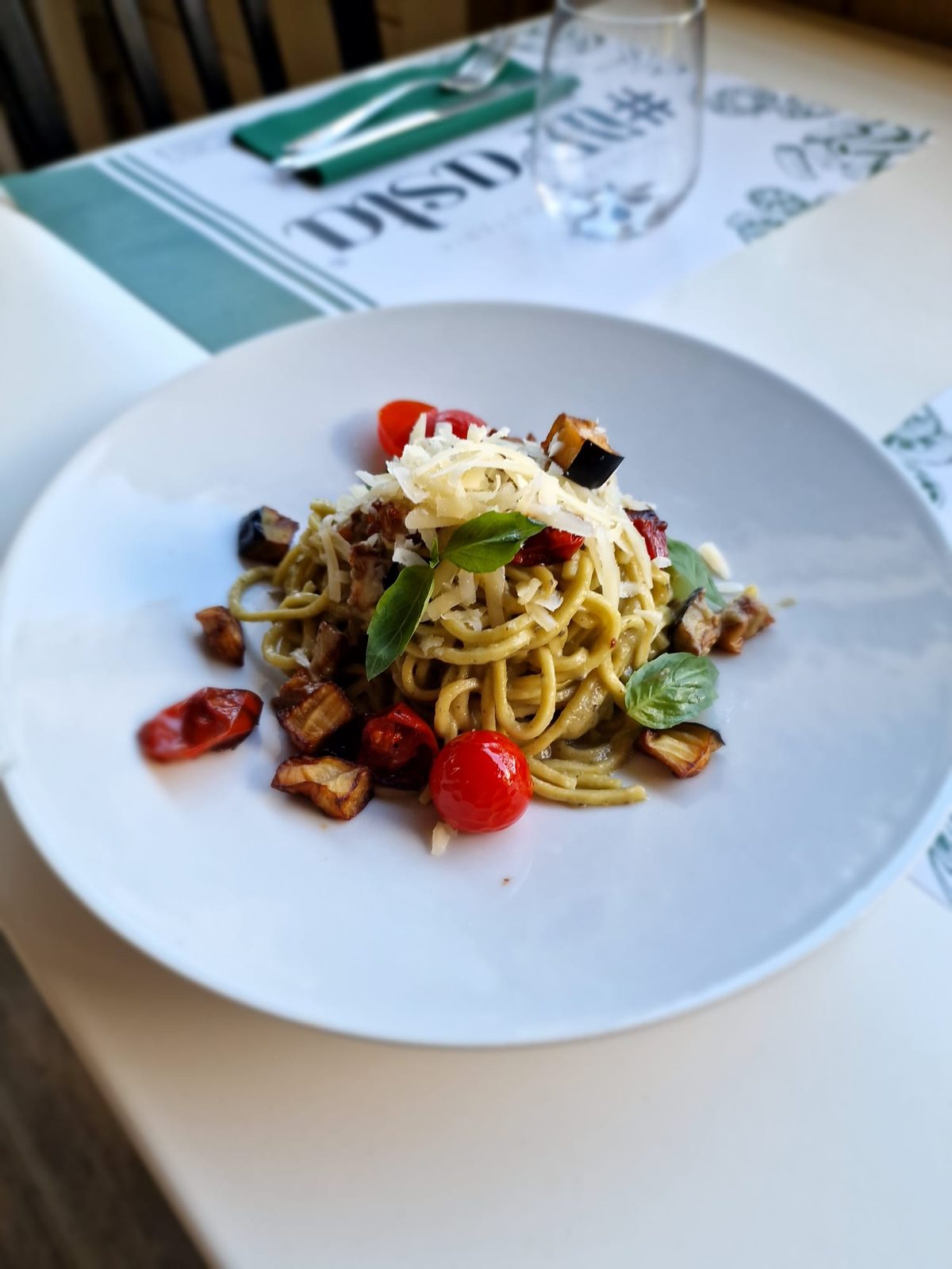 Spaghetti con crema di melanzane, pomodorini confit, basilico e cacio grattugiato, su un piatto bianco.