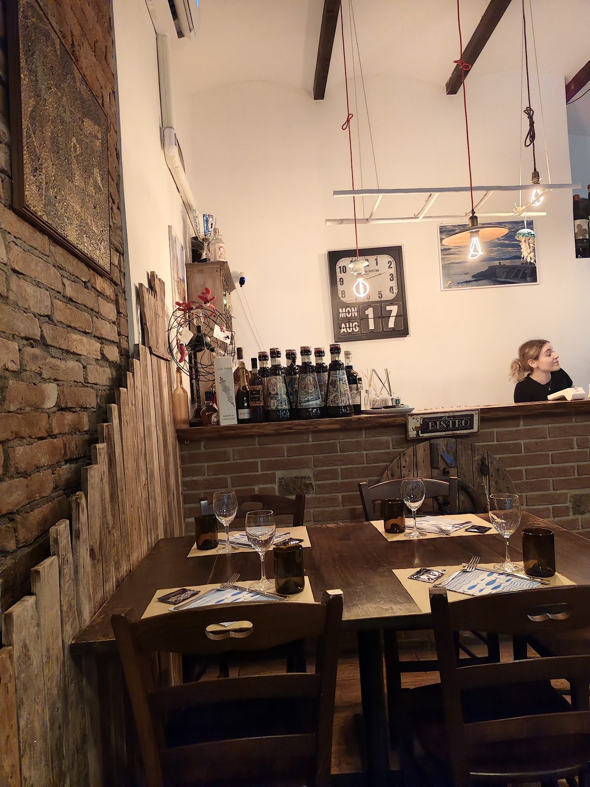 La sala del bistrot: muro di mattoni a vista, tavoli apparecchiati con calici e una mensola di bottiglie di vino sul fondo.
