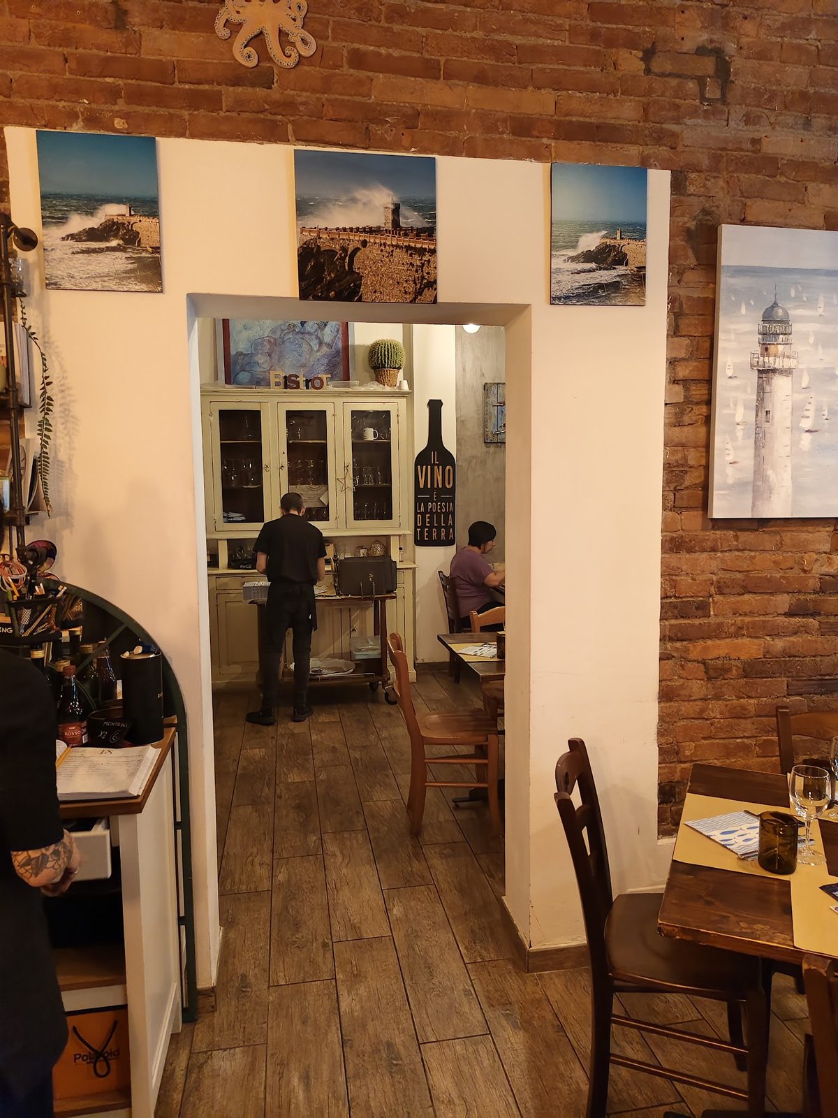 Vista dall&rsquo;ingresso verso la sala: un arco, le fotografie della costa appese al muro chiaro e i tavoli in fondo.